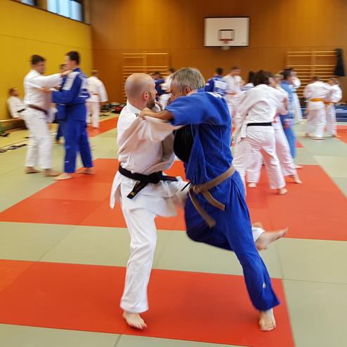 Zwei Judokas im Kampf auf einer Tatami, umgeben von weiteren Trainierenden in Judogis.