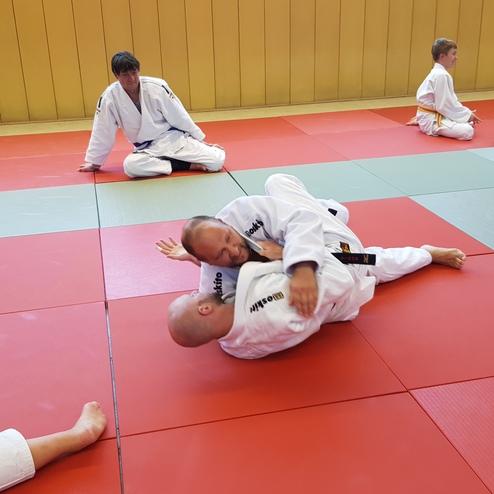 Zwei Personen im Judogi trainieren auf einer roten Matte, w&auml;hrend andere Teilnehmer im Hintergrund &uuml;ben oder sich ausruhen.