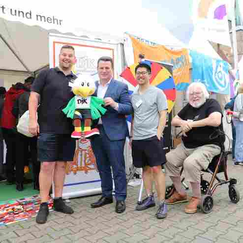 Vier Personen stehen vor einem Stand, halten eine Stofffigur und posieren l&auml;chelnd f&uuml;r das Foto. Bunte Banner im Hintergrund.