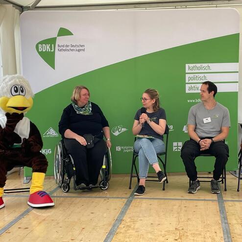 Diskussion auf einer B&uuml;hne: Vier Personen, davon eine mit Mobilit&auml;tshilfen, und ein Maskottchen, vor gr&uuml;ner Hintergrundwand.