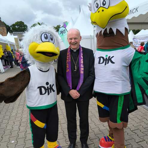 Drei Figuren stehen auf einer Veranstaltung: Zwei Maskottchen in Sporttrikots und ein Mann in schwarzer Kleidung mit lila Schal.