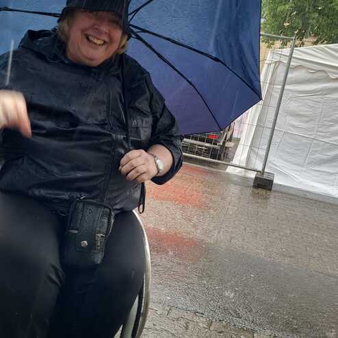 L&auml;chelnde Person im Rollstuhl, die unter einem blauen Regenschirm sitzt, w&auml;hrend es stark regnet.