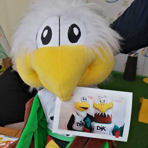 Pl&uuml;schmaskottchen eines Vogels h&auml;lt ein Foto von sich selbst in sportlicher Kleidung. Hintergrund mit Veranstaltungsbanner.