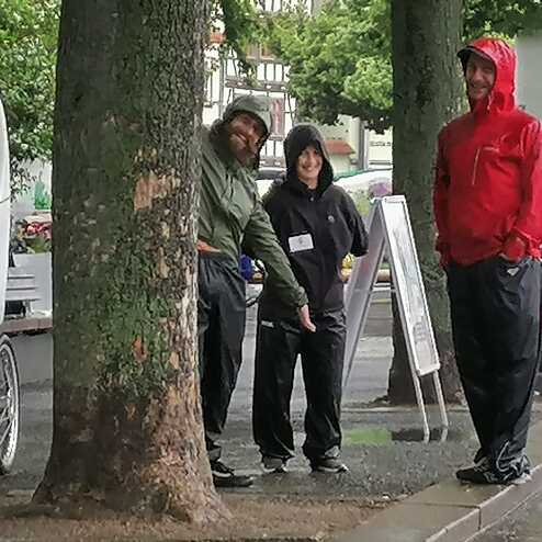 Drei Personen in Regenkleidung stehen an einem Stra&szlig;enrand unter B&auml;umen, w&auml;hrend es regnet. Ein Fahrzeug ist im Hintergrund sichtbar.