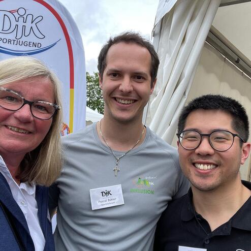 Drei Personen posieren lachend vor einem Banner der DJK Sportjugend, das im Hintergrund sichtbar ist.