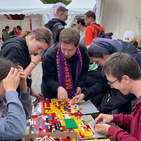 Sechs Personen bauen gemeinsam an einem bunten Lego-Projekt an einem Tisch im Freien.