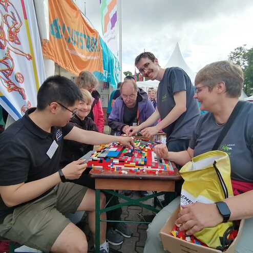 Sechs Personen bauen gemeinsam mit bunten LEGO-Steinen an einem Tisch, umgeben von Zelten und Bannern.