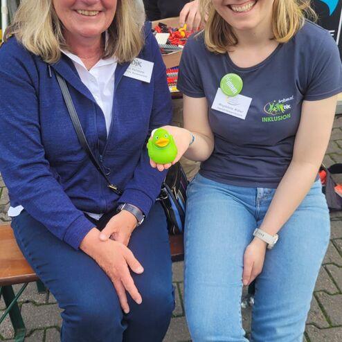 Zwei Frauen sitzen auf einer Bank, eine h&auml;lt eine gr&uuml;ne Gummiente, im Hintergrund ist eine Person mit Lego besch&auml;ftigt.