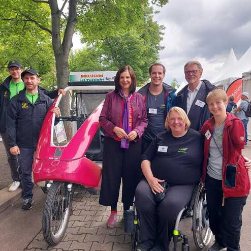 Gruppe von sechs Personen, darunter eine Rollstuhlfahrerin, vor einem pinkfarbenen Fahrrad in einem Park.