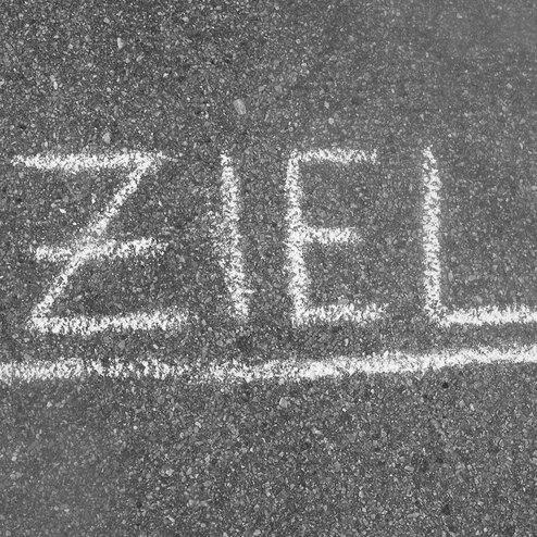 Wei&szlig;es Kreideschriftzug "ZIEL" auf einem grauen Asphaltboden.