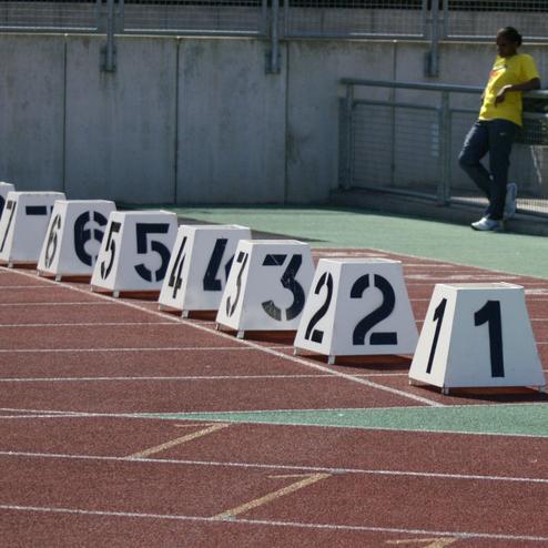 Startbl&ouml;cke mit Zahlen von 1 bis 8 auf einer roten Laufbahn, im Hintergrund steht eine Person in gelbem T-Shirt.