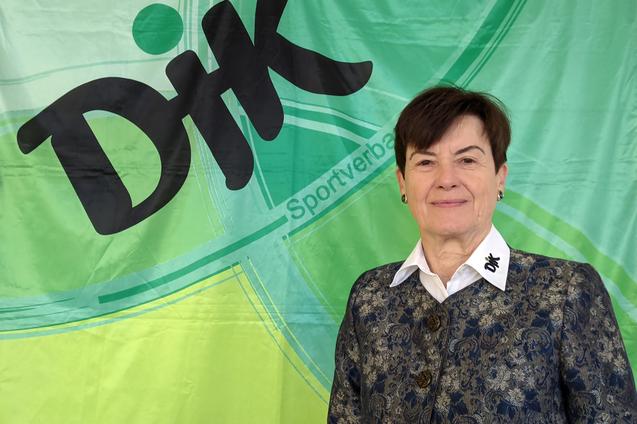 Frau mit k&uuml;rzeren Haaren, in einer eleganten Jacke, steht vor einem gr&uuml;nen Banner mit dem Schriftzug &bdquo;DiK Sportverband&ldquo;.