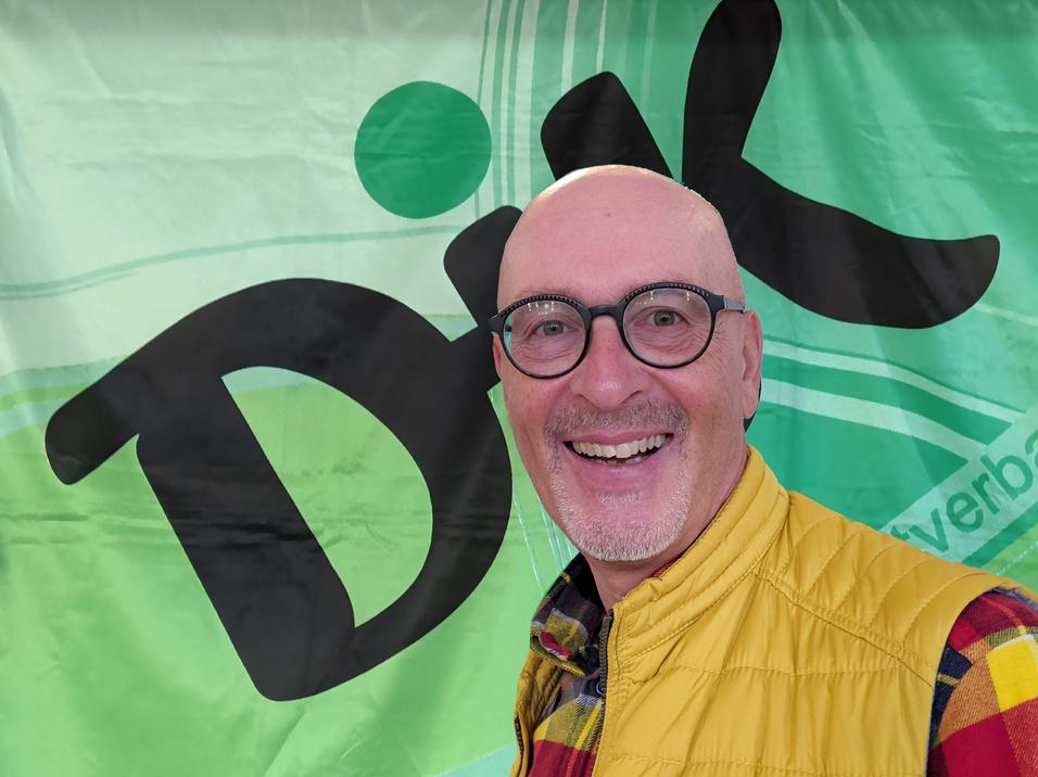 Mann mit Glatze und Brille l&auml;chelt vor einer bunten gr&uuml;nen Wand mit dem Schriftzug &bdquo;DiK&ldquo;.