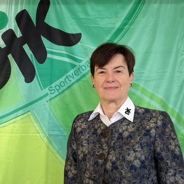 Frau mit k&uuml;rzeren Haaren, in einer eleganten Jacke, steht vor einem gr&uuml;nen Banner mit dem Schriftzug &bdquo;DiK Sportverband&ldquo;.
