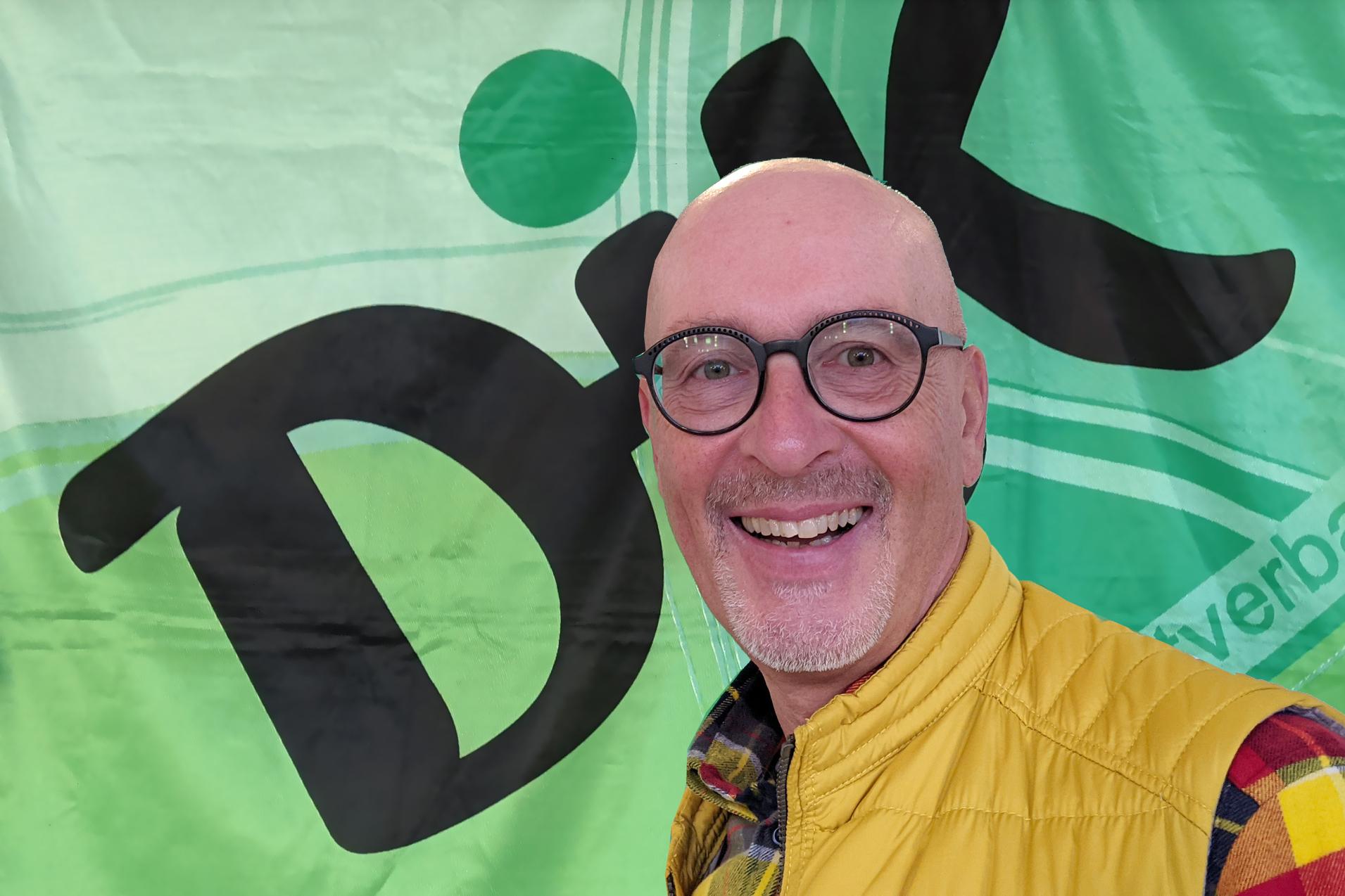 Mann mit Glatze und Brille l&auml;chelt vor einer bunten gr&uuml;nen Wand mit dem Schriftzug &bdquo;DiK&ldquo;.