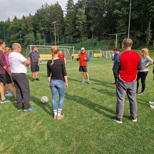 Gruppe von Personen um einen Trainer auf einem Fu&szlig;ballplatz, bereit f&uuml;r ein Training mit einem Ball im Mittelkreis.
