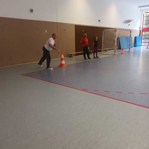 Ein Mann wirft einen Ball in einer Sporthalle, w&auml;hrend zwei Personen und ein Tor im Hintergrund stehen.