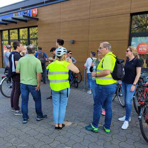 Gruppe von Radfahrern in Warnwesten steht vor einem Geb&auml;ude und diskutiert, Fahrr&auml;der sind daneben abgestellt.