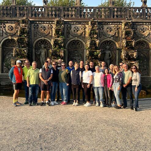 Gruppe von 20 Personen steht vor einer verzierten Wand mit Statuen und Pflanzen in einem sonnigen Garten.