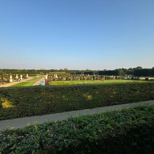 Weitl&auml;ufiger Garten mit Hecken, Skulpturen und Blumenbeeten unter blauem Himmel.