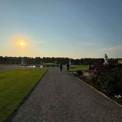 Paar spaziert auf einem Kiesweg im Garten, umgeben von Blumenbeeten, mit Sonnenuntergang im Hintergrund.