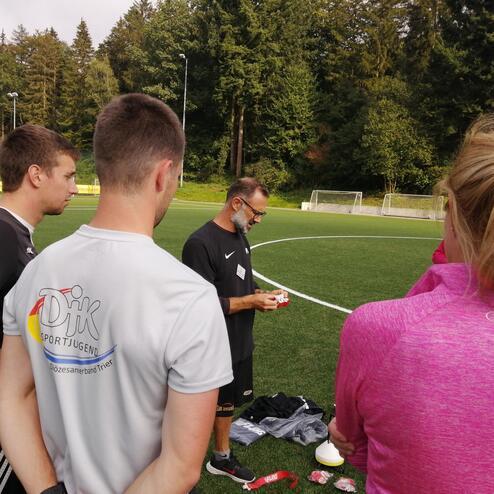 Trainer erkl&auml;rt einer Gruppe von Sportlern auf dem Fu&szlig;ballplatz eine &Uuml;bung, umgeben von B&auml;umen und Toren im Hintergrund.