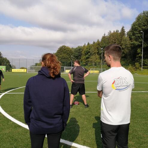 Zwei Personen beobachten Trainer auf einem Fu&szlig;ballplatz mit Kunstgrass und umgebender Natur.