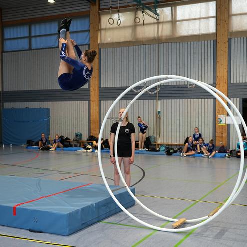 Sportlerin springt &uuml;ber zwei gro&szlig;e Ringe in einer Turnhalle, w&auml;hrend Zuschauer am Rand beobachten.