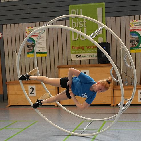 Akrobatin in blauer T-Shirt f&uuml;hrt eine &Uuml;bung im Rad in einer Sporthalle durch, w&auml;hrend sie auf einer Seite liegt.