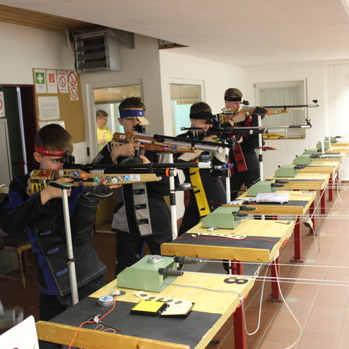 Sechs Jugendliche stehen an Tischtennistisch mit Luftgewehren und zielen auf Zielscheiben in einem &Uuml;bungsraum.