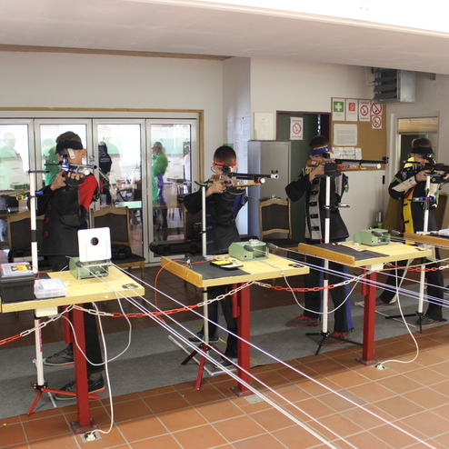 Sechs Sch&uuml;tzen in Wettkampfkleidung zielen mit Luftgewehren auf entsprechende Zielscheiben an Holztischen.