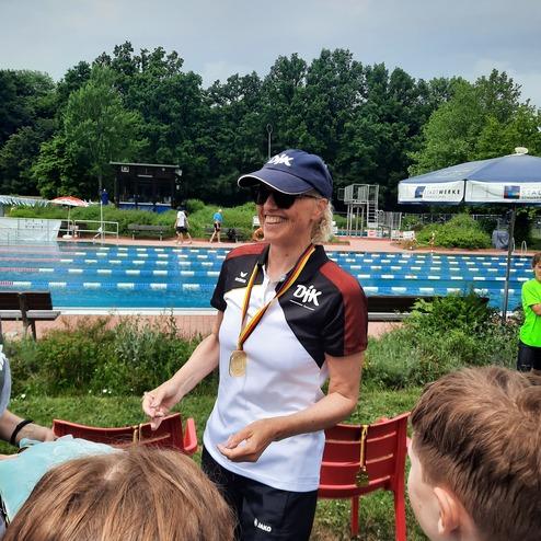 Frau mit Medaille steht l&auml;chelnd am Schwimmbecken, umgeben von Kindern und Sportausr&uuml;stung.