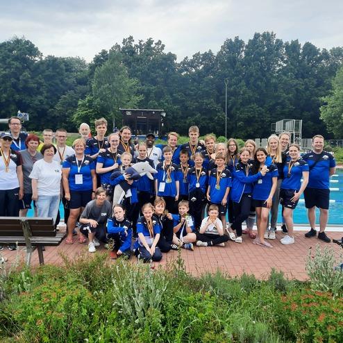 Gruppenshot von Schwimmern und Trainern in blauen Shirts, vor einem Schwimmbecken mit Medaillen und Pokalen.