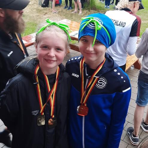 Zwei M&auml;dchen mit Medaillen stehen l&auml;chelnd unter einem Baum, umgeben von Zuschauern bei einer Sportveranstaltung.