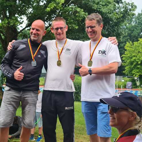 Drei Medaillentr&auml;ger posieren l&auml;chelnd auf einer Wiese, umgeben von Zuschauern und B&auml;umen im Hintergrund.