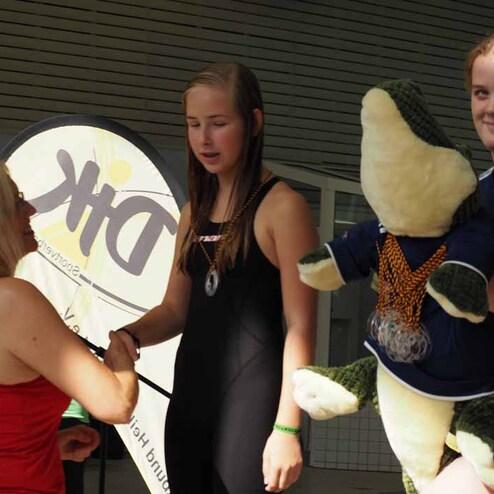 Zwei Schwimmerinnen erhalten eine Auszeichnung, eine sch&uuml;ttelt die Hand einer Frau, im Hintergrund ein gro&szlig;es Schild.