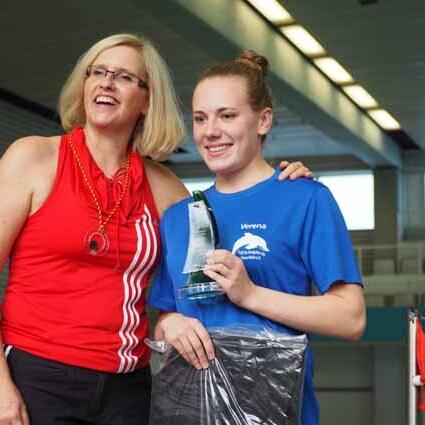 Zwei Frauen posieren l&auml;chelnd bei einer Siegerehrung; eine h&auml;lt einen Pokal, die andere steht daneben.