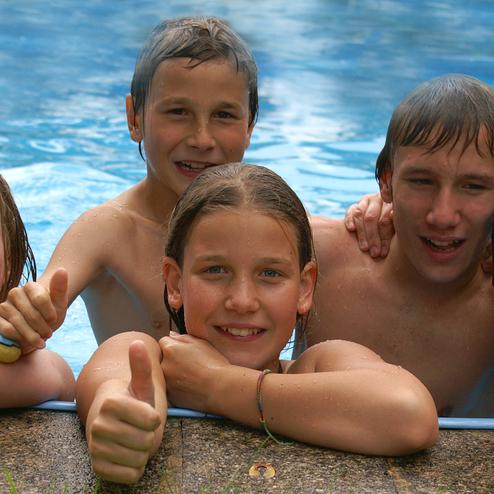 F&uuml;nf Kinder im Wasser zeigen lachend Daumen hoch, w&auml;hrend sie am Beckenrand posieren.