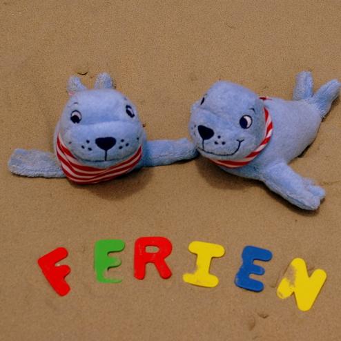 Zwei blaue Kuschelrobben auf Sand, umgeben von bunten Buchstaben, die &bdquo;FERIEN&ldquo; bilden.
