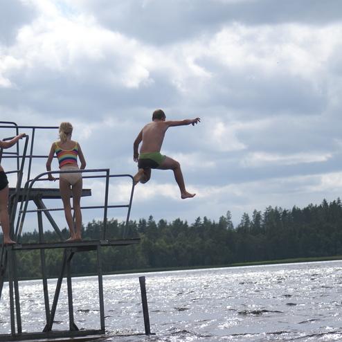 Drei Kinder springen von einem Sprungturm ins Wasser eines Sees unter einem bew&ouml;lkten Himmel.