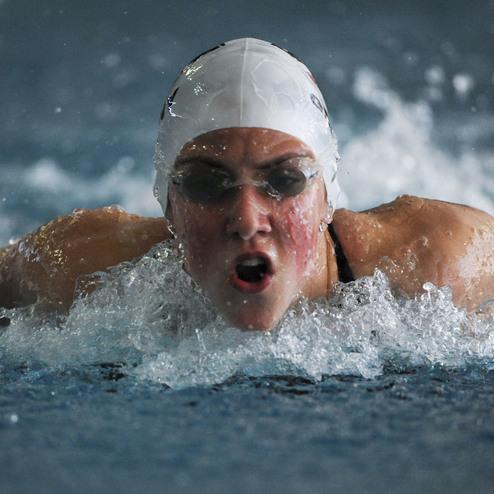 Schwimmerin in einem Wettkampf mit intensivem Gesichtsausdruck, der Kopf &uuml;ber Wasser, spritzendes Wasser um sie herum.