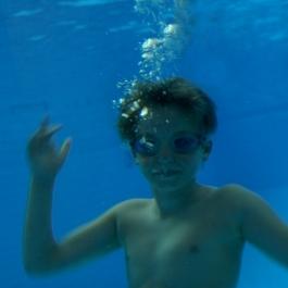 Junge mit Schwimmbrille unter Wasser, zeigt die H&auml;nde nach oben, umgeben von blauen Wasserblasen.