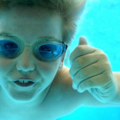 Junge mit blonden Haaren und Schwimmbrille taucht unter Wasser und zeigt Daumen hoch.