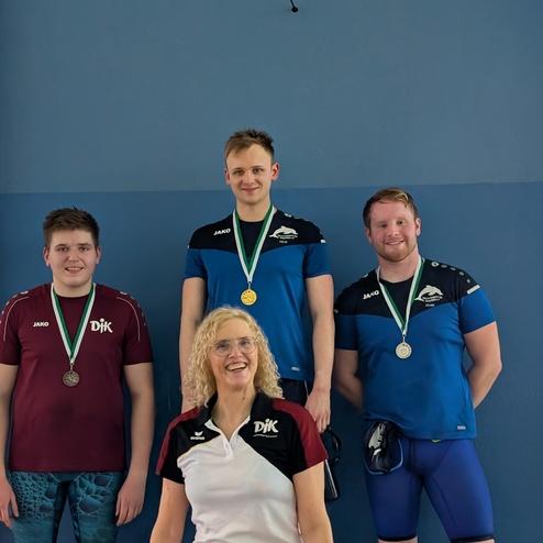 Drei m&auml;nnliche Sportler und eine Trainerin stehen vor einer blauen Wand, alle tragen Medaillen und Sportbekleidung.