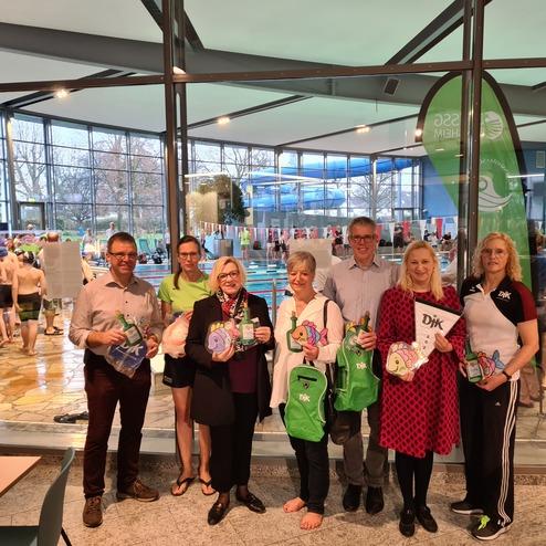 Gruppe von sieben Personen in Schwimmbad mit Geschenken und Taschen, im Hintergrund Schwimmer und Wassersportbereich sichtbar.
