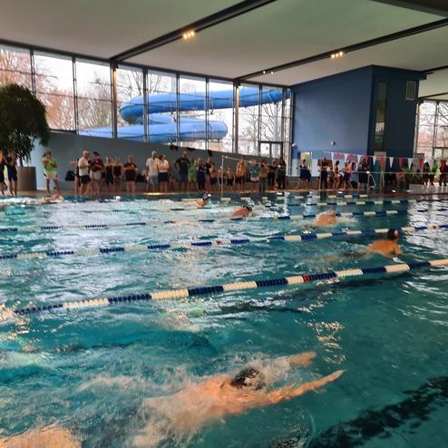Wettkampf im Schwimmbad mit mehreren Schwimmern und zuschauenden Personen an der Beckenrand. Rutschbahn im Hintergrund.