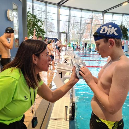 Trainerin spricht mit einem jungen Schwimmer am Beckenrand eines Schwimmbads, im Hintergrund sind weitere Schwimmer.