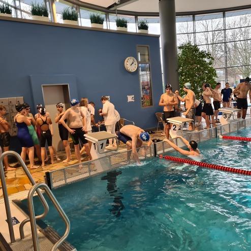 Schwimmer in Badekleidung stehen an Startbl&ouml;cken entlang eines Schwimmbeckens in einem modernen Sportbad.