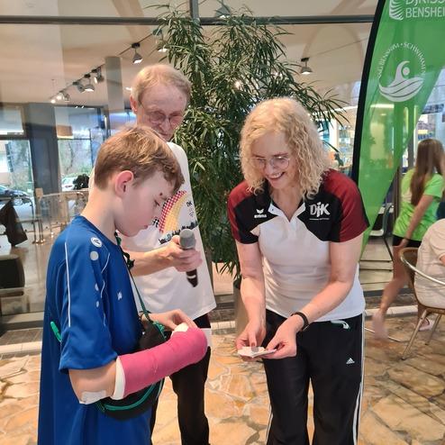 Drei Personen stehen in einem Raum, zwei Erwachsenen und einem Jungen mit Gipsarm, der ein Handy zeigt.