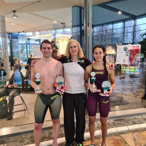 Drei Menschen stehen in einem Schwimmbad: zwei Sportler mit Pokalen und eine Trainerin in sportlichem Outfit.